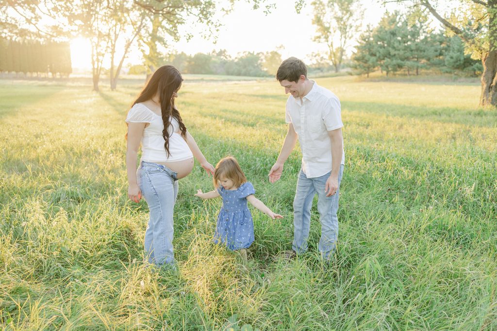 A pregnant couple playfully interacts with their toddler in an open field in Minnesota with the setting sun casting a golden glow throughout the image.