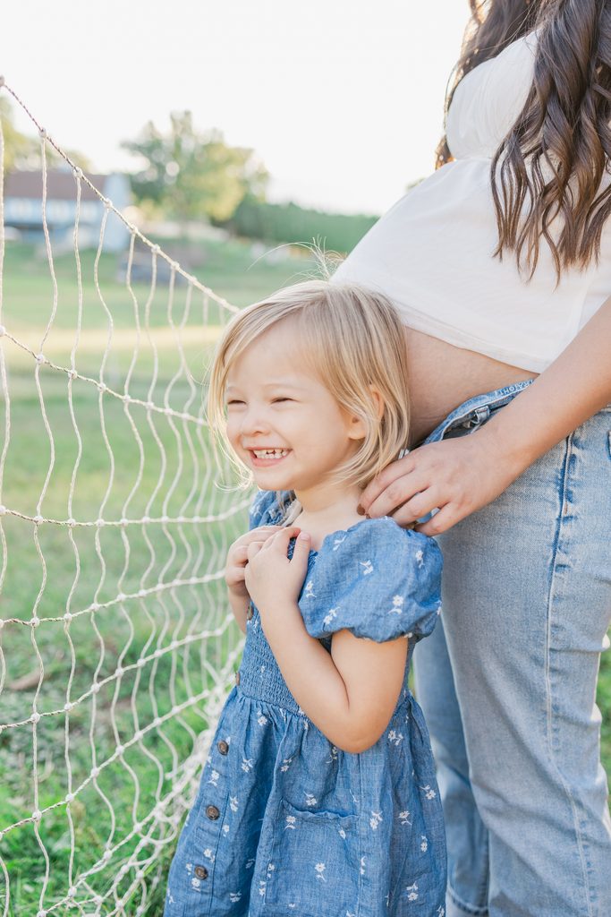 A smiling toddler stands in front of a pregnant mother with the setting sun glowing behind them during a maternity session outdoors near Woodbury, MN.