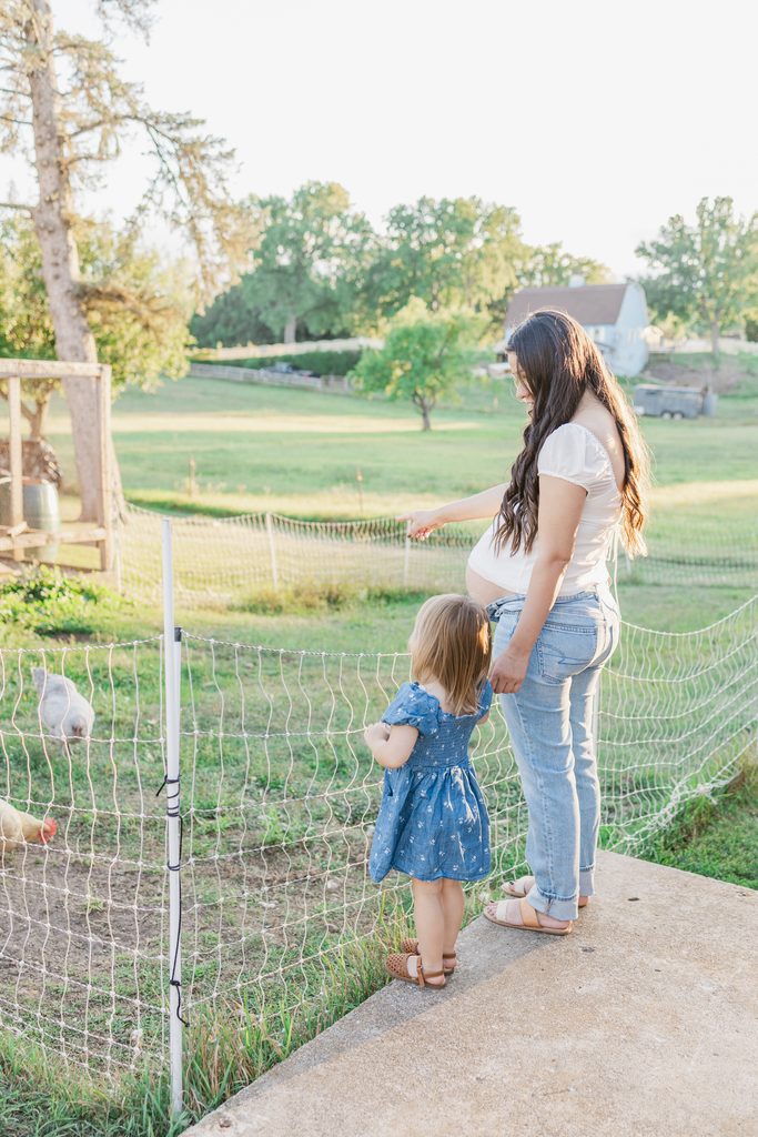 Pregnant mother stands with her toddler at a farm, looking out at a chicken coop during a lifestyle maternity photo session in Afton, MN.