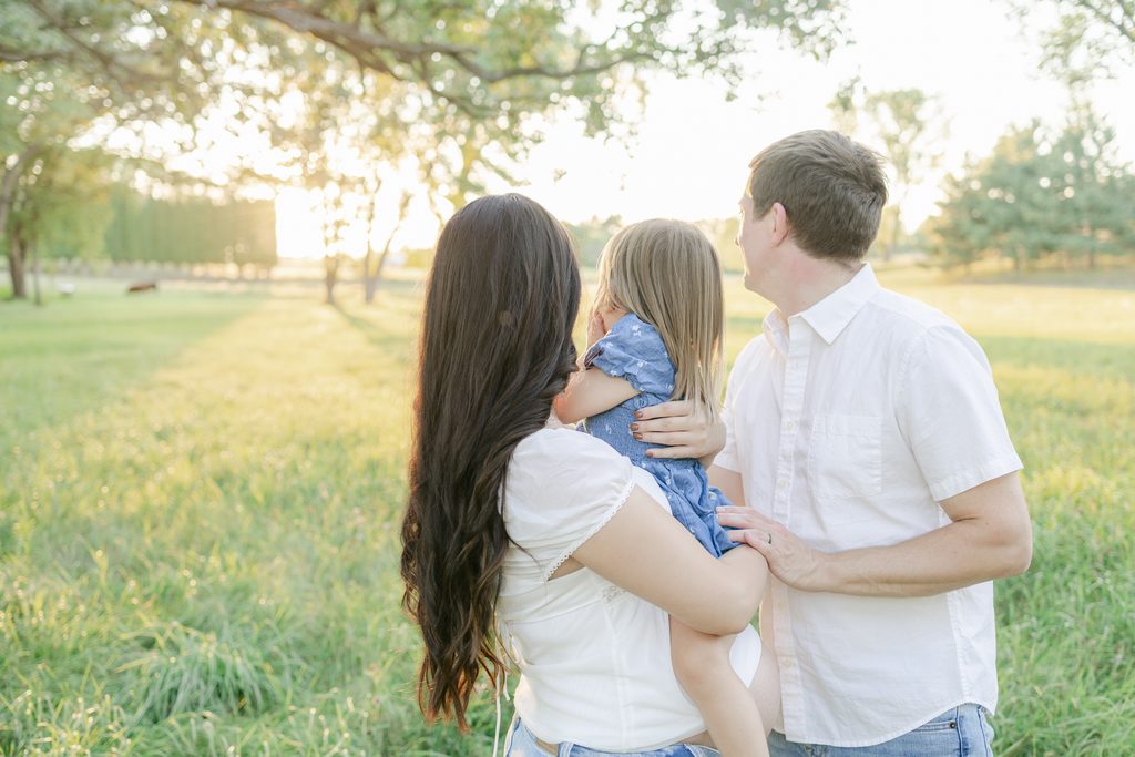A couple and their toddler snuggle as they gaze away from the camera at the setting sun and a field of cows near Woodbury, MN.